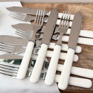 May include: A collection of stainless steel forks and knives with white handles. The silverware is arranged on a wooden surface and a white cloth. The knives have a curved blade and the forks have three tines. The silverware is for dining.