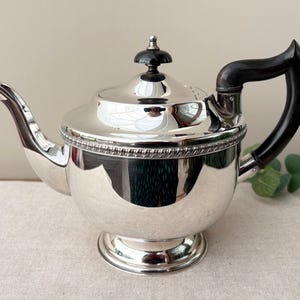 May include: A polished silver teapot featuring a rounded body, a curved spout, and a dark black handle. The lid is topped with a small black finial, and the upper rim of the teapot is decorated with a detailed beaded border design.