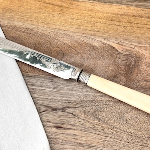 Puede incluir: Un cuchillo de mantequilla plateado con mango color marfil. La hoja tiene un diseño floral ornamentado. El cuchillo está sobre una superficie de madera junto a un paño blanco.