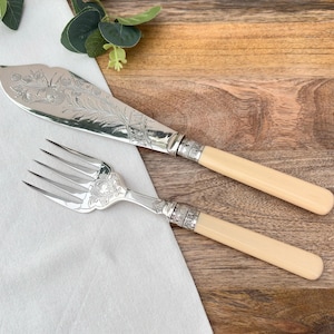 May include: Silver butter knife and fork set with cream-coloured handles. The knife blade and fork tines are silver with ornate floral and leaf engravings. The handles are a light cream colour, placed on a white cloth and wooden surface.