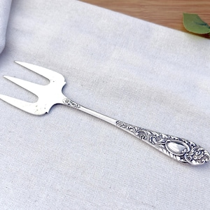 May include: A silver three-pronged fork with an ornate handle. The fork is on a light blue linen cloth. The handle has an oval design and detailed floral patterns. The tines are straight and evenly spaced.