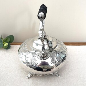 May include: A silver-plated tea kettle featuring an ornate embossed floral pattern across its rounded body. The kettle stands on three decorative feet and includes a hinged lid with a central handle topped by a black cylindrical grip.