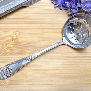 May include: A silver serving spoon with a decorative handle and a perforated bowl. The spoon is on a wooden surface.