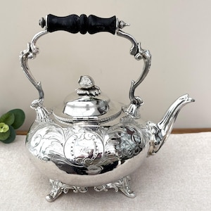 May include: A silver-plated tea kettle featuring intricate embossed floral scrollwork, a hinged lid with a decorative finial, and a black wooden handle. The kettle stands on four ornate feet and includes a curved spout and a central engraved cartouche.