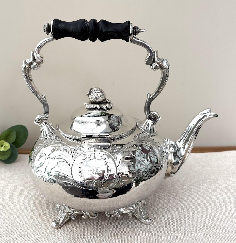 May include: A silver-plated tea kettle featuring intricate embossed floral scrollwork, a hinged lid with a decorative finial, and a black wooden handle. The kettle stands on four ornate feet and includes a curved spout and a central engraved cartouche.