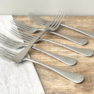 May include: A set of five silver-toned metal forks arranged in a fan-like pattern. Each fork features four tines and a smooth, rounded handle. The forks have a polished finish and are positioned overlapping one another, showcasing their uniform design.