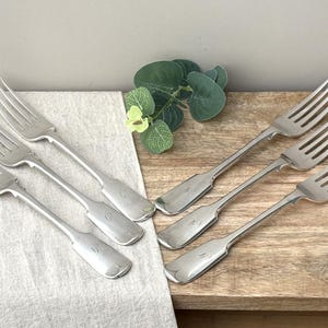 May include: A set of six silver-toned metal forks with four tines and a smooth, flat handle design. The forks are arranged in two groups, with three forks resting on a light-coloured fabric cloth and three forks resting on a wooden surface.