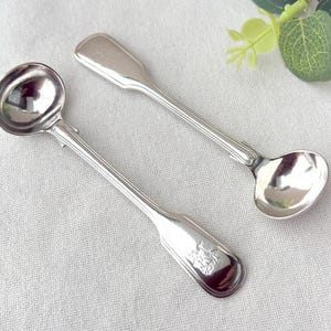 May include: Two silver-toned metal spoons with round bowls and long, slender handles. The handles feature a ribbed design and a small engraved crest or emblem at the end. The spoons have a polished, reflective finish and a classic, symmetrical shape.