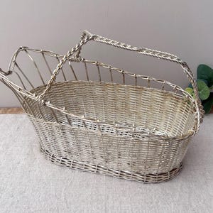 May include: A silver-toned metal wine bottle cradle featuring a woven wire basket design with an oval base, an open top, and a single twisted metal handle that extends across the top to facilitate pouring from a bottle placed inside the frame.
