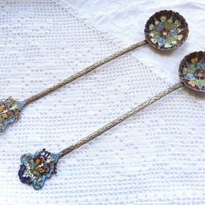 May include: A pair of antique silver spoons with colorful enamel floral designs. The spoons have long handles and ornate details.