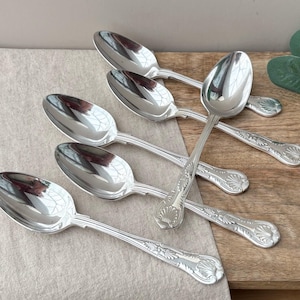 May include: A set of five silver-toned metal spoons with polished oval bowls and handles featuring a decorative embossed shell motif at the base. The spoons are arranged in a fan-like pattern, showcasing the reflective metallic finish and the detailed scrollwork on the handles of each individual piece.