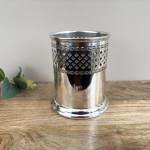 Puede incluir: Una taza de metal plateado con una banda decorativa con diseños de diamantes y flores. La taza tiene una base acampanada y una superficie lisa y reflectante. El borde superior también es liso y brillante.