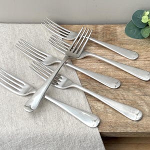 May include: A set of five silver-toned stainless steel forks with four tines each. The forks feature a smooth, polished finish with a rounded handle design. They are arranged in a overlapping pattern, showcasing the uniform shape and metallic luster.