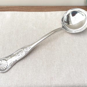 May include: A polished silver-toned metal soup ladle featuring a deep, circular bowl and a long, slender handle. The handle is decorated with an embossed shell motif at the base and intricate scrollwork patterns extending along the stem.