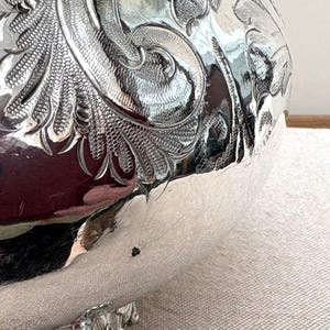 May include: A polished silver bowl featuring intricate, deeply engraved scrollwork and floral patterns across its curved surface. The metallic finish is highly reflective, showing distorted surroundings and a small dark spot on the lower side.