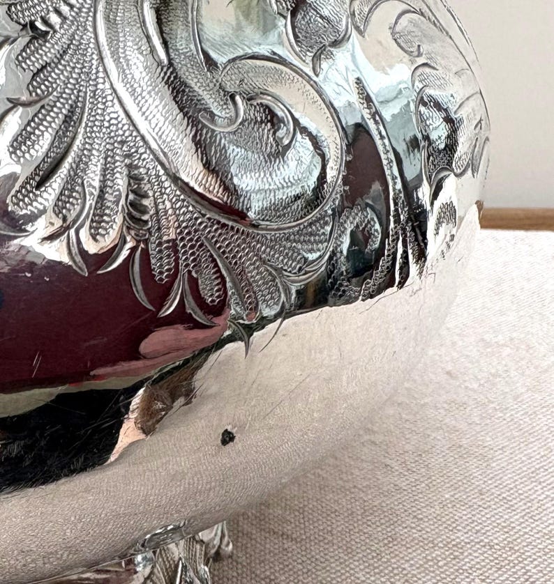 May include: A polished silver bowl featuring intricate, deeply engraved scrollwork and floral patterns across its curved surface. The metallic finish is highly reflective, showing distorted surroundings and a small dark spot on the lower side.