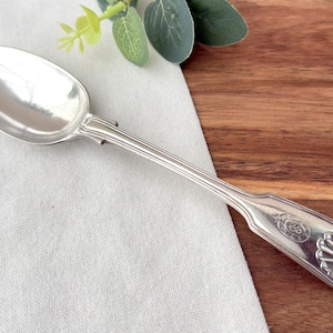 May include: A silver spoon with an oval bowl and a decorative handle. The handle has vertical lines and a shell-shaped design. The spoon is on a white cloth. The spoon is made of silver.