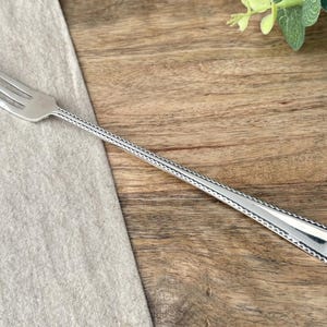 May include: A silver-toned metal cocktail fork featuring three slender tines and a long, narrow handle. The handle is decorated with a repeating embossed zigzag or scalloped pattern along its edges, extending from the base of the tines to the end.