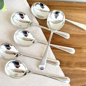 May include: A set of five polished silver-tone metal soup spoons with rounded bowls and smooth, plain handles. The spoons are arranged in a cluster, reflecting light on their shiny surfaces, showcasing a simple and functional design for dining.