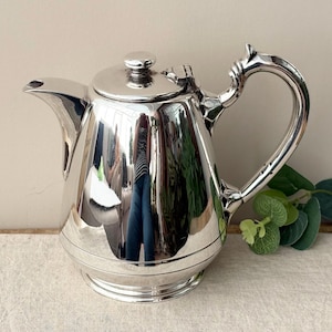 May include: A polished silver-toned metal teapot featuring a wide, rounded base, a tapered body, and a curved handle with decorative detailing. The teapot includes a hinged lid with a small knob and a short, angled spout, reflecting its surroundings.