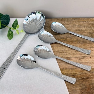 Puede incluir: Un juego de cinco cucharas plateadas con un diseño floral en el cuenco y un mango texturizado. Las cucharas están dispuestas sobre una superficie de madera con un paño blanco y hojas verdes en el fondo. Las cucharas varían en tamaño.