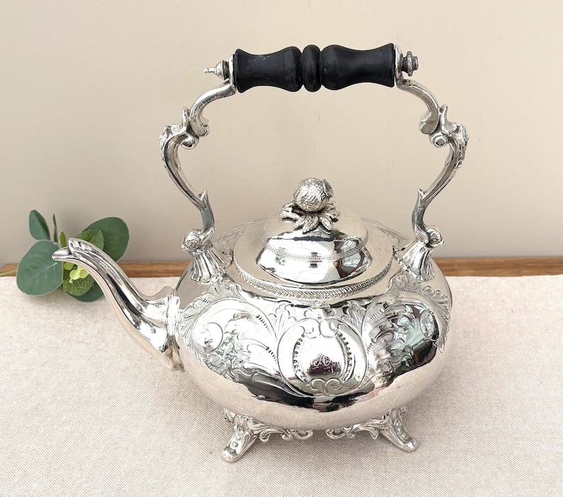 May include: A silver-plated tea kettle featuring intricate floral engravings on the body, a curved spout, and a black wooden handle. The lid is topped with a decorative finial, and the base rests on four ornate feet with detailed scrollwork.