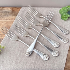May include: A set of six silver-toned metal forks arranged in a fan pattern. Each fork features four tines and a handle decorated with a raised shell motif at the base. The polished metal surfaces reflect light, highlighting the smooth curves.