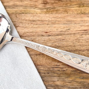 May include: A silver ladle with an oval bowl and a long, decorated handle. The handle features ornate floral and geometric patterns. The ladle rests on a white cloth against a wooden background.