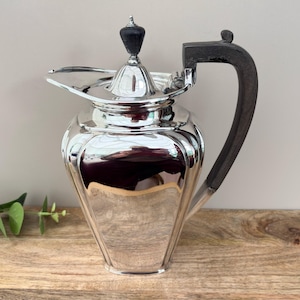 May include: A silver pitcher with a black handle and lid finial. The pitcher has a geometric design and a spout. The handle is curved and attached to the body. The pitcher is likely made of silver-plated metal.