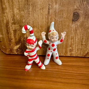 May include: Two vintage ceramic figurines of elves. One elf is wearing a red and white striped outfit with a matching hat. The other elf is wearing a white outfit with red polka dots and a matching hat. Both elves have their arms raised.