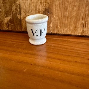 May include: A small, white ceramic egg cup with a crackled glaze. The cup has a flared rim and a pedestal base. The letters "V.F" are printed in black on the front. The egg cup is on a wooden surface.