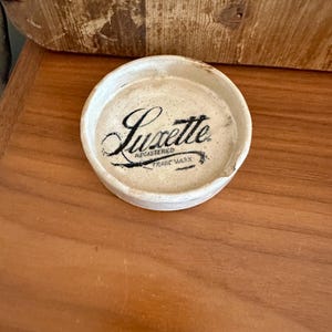 May include: A small, round, off-white ceramic lid with the word "Luxette" in black script. The text "Registered Trade Mark" is below the name. The lid has a chipped edge and sits on a wooden surface.