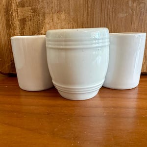 May include: Three white ceramic cups of varying shapes are arranged on a wooden surface. The central cup has a rounded body with horizontal ridges. The other two cups are cylindrical. The background features a wooden panel.