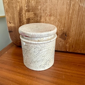 May include: A small, off-white ceramic jar with a lid. The jar has a crackle glaze pattern and a round shape. The lid is also off-white with a pinkish hue. The jar is sitting on a wooden surface.