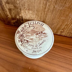 May include: A round, off-white container with a brown landscape design and text. The lid reads "Icilma Creme de Savon Pour la Barbe" in brown script. The container sits on a wooden surface.