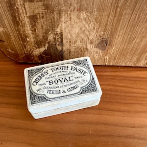 May include: A small, rectangular box of "Cherry Tooth Paste" with text in black and white. The box is on a wooden surface with a wooden background. The text on the box reads "Trade BOVAL Mark for Cleansing & Preserving the Teeth & Cums."