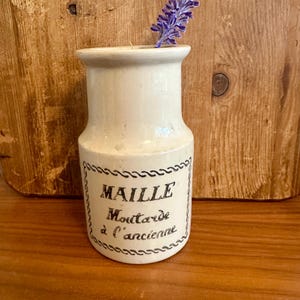 May include: A small, off-white ceramic jar with a label that reads "MAILLE Moutarde à l'ancienne" in black script. A sprig of purple lavender is placed inside the jar. The jar is set against a wooden background.