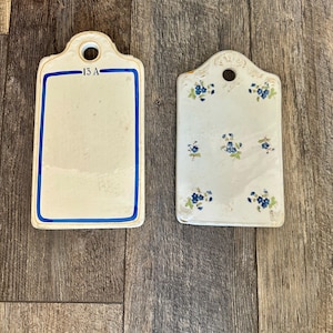 May include: Two vintage rectangular ceramic tags. One is white with a blue border and the text "15 A". The other is white with blue floral designs. Both have a hole at the top for hanging.