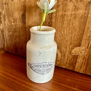 May include: A white ceramic vase with a crackled glaze and black lettering that reads "Poultons & Noel Home-Made Potted Meat Belgravian Brand" holds a single white felt flower with green felt leaves.