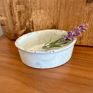 May include: A white ceramic bowl with a speckled glaze. The bowl is sitting on a wooden surface and has a sprig of lavender flowers inside.