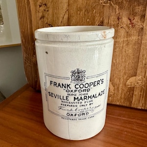 May include: A vintage, off-white ceramic jar of Frank Cooper's Seville Marmalade. The jar has a cylindrical shape with a slightly cracked surface. The label features black text and a crest, with the words "Oxford" and "Registered Trade Mark" visible.
