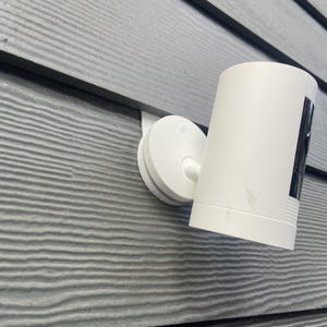 May include: A white security camera mounted on a gray siding wall. The camera is pointed downwards and has a black lens.