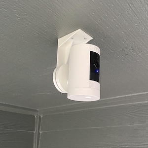 May include: A white Ring security camera mounted on a white bracket attached to a ceiling. The camera is pointed downwards.