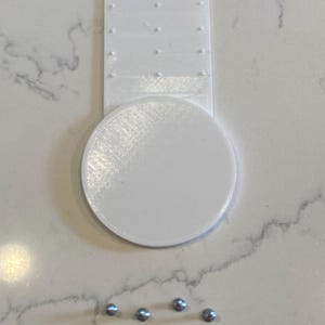May include: A white 3D-printed object with a circular base and a rectangular handle featuring small, evenly spaced holes. Four small, silver-colored metal balls are arranged below the object on a light-colored surface. The object appears to be a tool or a part of a larger device.