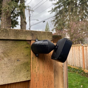 May include: A black security camera mounted on a wooden fence. The camera is pointed towards the ground and has a wide-angle lens.