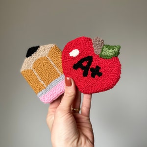 May include: A hand holds two felt ornaments, a red apple with a black "A+" and a yellow and pink pencil.