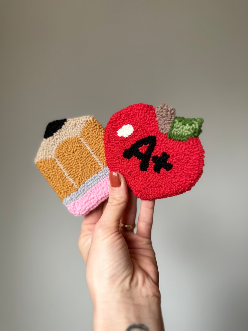 May include: A hand holds two felt ornaments, a red apple with a black "A+" and a yellow and pink pencil.