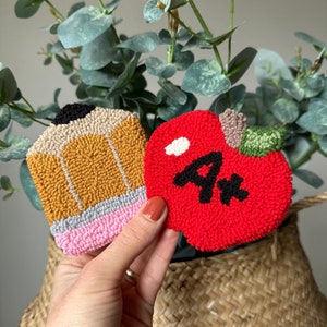 May include: Two felt crafts, a red apple with a black "4*" and a yellow pencil with a pink eraser, are held in a hand against a woven basket background.