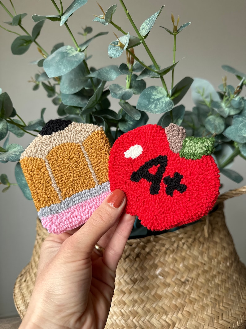 May include: Two felt crafts, a red apple with a black "4*" and a yellow pencil with a pink eraser, are held in a hand against a woven basket background.