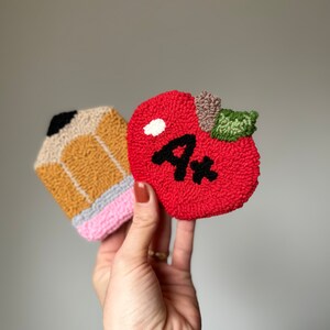 May include: A red felt apple with a green stem and a black "A+" on it. A yellow and brown felt pencil with a pink eraser is next to the apple.
